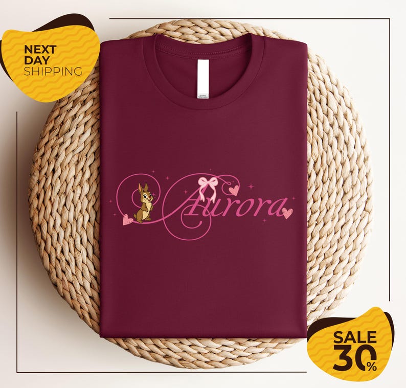 a burgundy t-shirt with a pink and purple design, including a cartoon character and the word "Aurora". The t-shirt is displayed on a woven basket, and there are two yellow sale signs in the background.