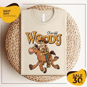 A t-shirt featuring a cartoon image of Woody, a character from the Toy Story franchise, riding a horse. The t-shirt is displayed on a woven basket, and there are two yellow sale signs indicating a 30% discount on the item.
