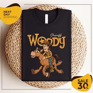 A black t-shirt with a cartoon image of Woody, a character from the Toy Story franchise, riding a brown horse. The t-shirt is displayed on a woven basket, and there are yellow price tags indicating a sale of 30% off.