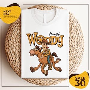 A white t-shirt with a cartoon image of Woody, a character from the Toy Story franchise, riding a brown horse. The t-shirt is displayed on a woven placemat, and there are yellow price tags indicating a sale of 30% off.