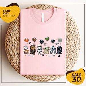 A pink t-shirt with a graphic design featuring various Star Wars characters, including Chewbacca, Stormtrooper, Darth Vader, and Baby Yoda, along with other Star Wars-themed elements.