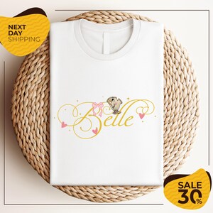 a white t-shirt with a printed design, including the word "Belle" and decorative elements such as hearts and a bow. The t-shirt is displayed on a woven basket, and there are two yellow sale signs in the image, one on the left and one on the right, indicating a sale or discount.