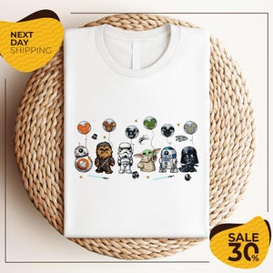 A white t-shirt with a graphic design featuring various Star Wars characters, including Chewbacca, Stormtrooper, Darth Vader, and Baby Yoda, along with other Star Wars-themed elements.
