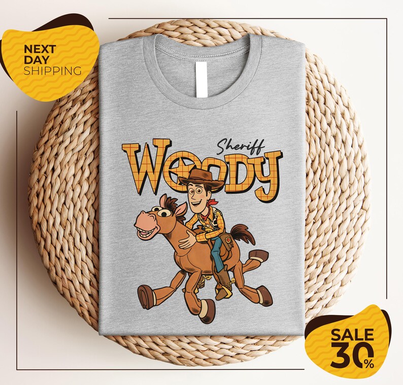 A gray t-shirt with a cartoon image of Woody, a character from the Toy Story franchise, riding a brown horse. The t-shirt is displayed on a woven basket, and there are yellow sale signs indicating a 30% discount.