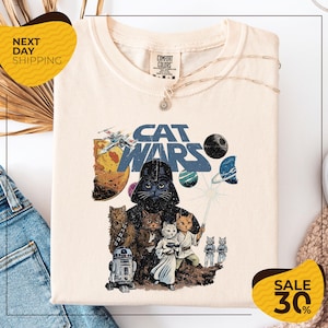 May include: Cream-colored t-shirt featuring a "Cat Wars" graphic. The design includes a cat version of Darth Vader, other cats as characters, and space-themed elements. The shirt has a crew neck and short sleeves.