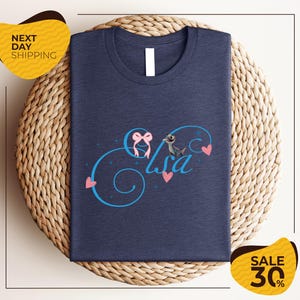 A navy blue t-shirt with a personalized name and heart design, placed on a woven placemat. The t-shirt is displayed for sale, with a "Next Day Shipping" label and a "Sale 30%" discount label.