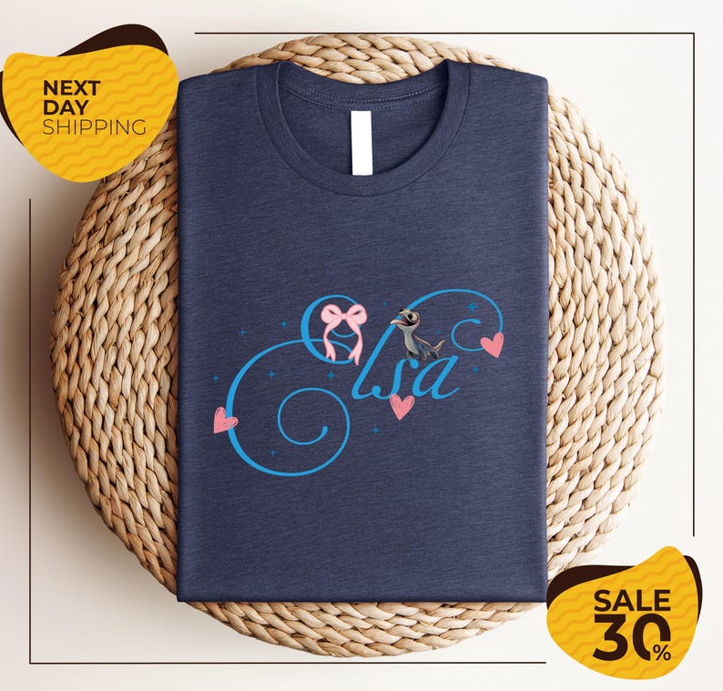 A navy blue t-shirt with a personalized name and heart design, placed on a woven placemat. The t-shirt is displayed for sale, with a "Next Day Shipping" label and a "Sale 30%" discount label.