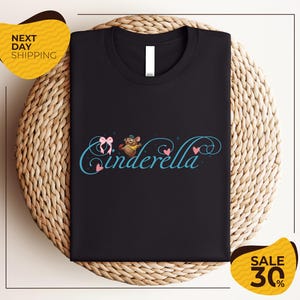 a black t-shirt with a whimsical design, including a character resembling Cinderella, placed on a woven placemat. The t-shirt is displayed on a table, and there are two yellow sale signs in the background, indicating a sale or discount.