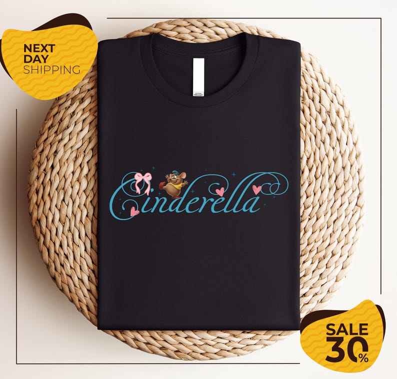 a black t-shirt with a whimsical design, including a character resembling Cinderella, placed on a woven placemat. The t-shirt is displayed on a table, and there are two yellow sale signs in the background, indicating a sale or discount.