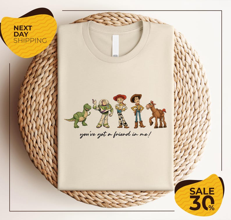 a beige t-shirt with a graphic design of five cartoon characters from the Toy Story franchise, including Woody, Buzz Lightyear, Timmy, and a horse. The text below the characters reads "you've got a friend in me!"