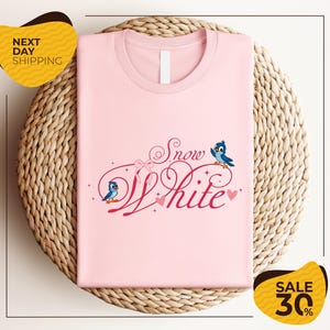 A pink t-shirt with the text "Snow White" and two blue birds, placed on a woven basket.