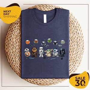 A blue t-shirt with a graphic design featuring various Star Wars characters, including Chewbacca, Stormtrooper, Baby Yoda, and Darth Vader, arranged in a row.