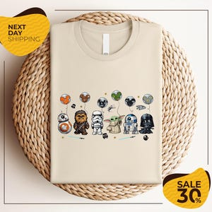 a beige t-shirt with a graphic design of various Star Wars characters, including Chewbacca, Stormtrooper, Darth Vader, and Baby Yoda, along with other Star Wars-themed elements.