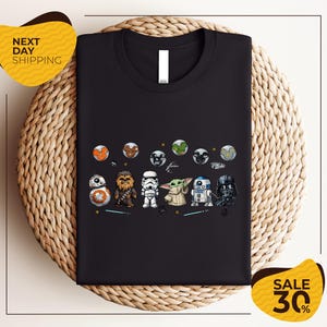 a black t-shirt with a graphic design of various Star Wars characters, including the main characters from the Star Wars franchise, such as Rey, Finn, and Chewbacca, along with other iconic characters like Bond, Baby Yoda, and Darth Vader. The t-shirt is displayed on a woven placemat, and there are two yellow sale signs in the image, indicating a discount or sale event.