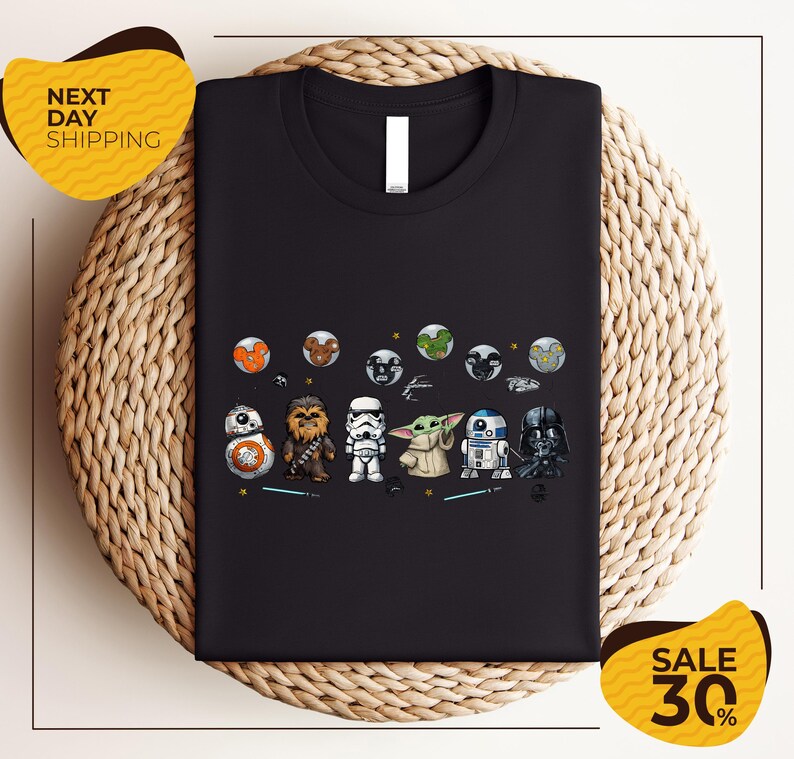a black t-shirt with a graphic design of various Star Wars characters, including the main characters from the Star Wars franchise, such as Rey, Finn, and Chewbacca, along with other iconic characters like Bond, Baby Yoda, and Darth Vader. The t-shirt is displayed on a woven placemat, and there are two yellow sale signs in the image, indicating a discount or sale event.