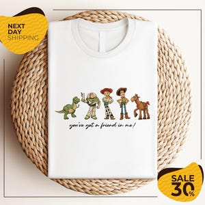 A white t-shirt with a graphic design featuring characters from the Toy Story movie, including Woody, Buzz Lightyear, and a horse. The text below the characters says "you've got a friend in me!"