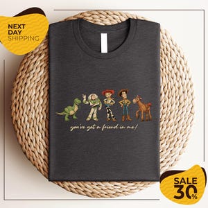 a gray t-shirt with a graphic design of characters from the Toy Story movie, including Woody, Buzz Lightyear, and a dinosaur. The text on the t-shirt says "you've got a friend in me!"