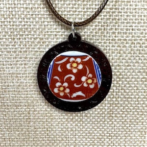 May include: A circular pendant necklace featuring a dark brown wooden frame with an intricate carved border. The center inlay displays a red ceramic piece decorated with white floral patterns, yellow centers, and blue striped side accents.