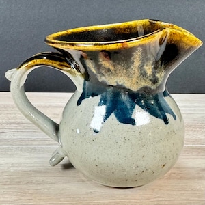 May include: A ceramic pitcher with a rounded body and a curved handle. The base is a speckled light gray, transitioning into a deep blue drip pattern, while the rim and upper section feature a glossy, toasted golden-brown glaze finish.