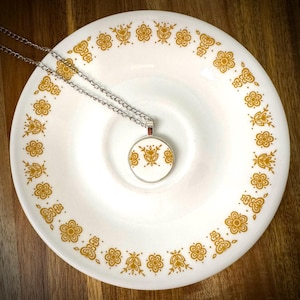 May include: A circular pendant necklace featuring a white ceramic base with a gold-colored floral pattern, suspended from a silver-toned metal chain. The pendant design matches the repeating gold flower motif printed along the rim of the plate.