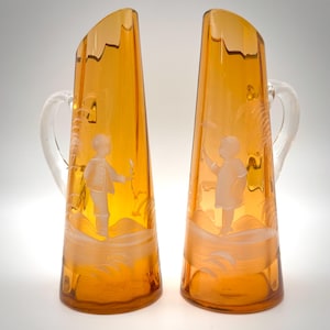 May include: Two tall, amber-colored glass pitchers with clear glass handles. Each pitcher features a slanted top rim and an etched, frosted white design depicting a figure standing in a landscape with foliage, set against a smooth, translucent body.
