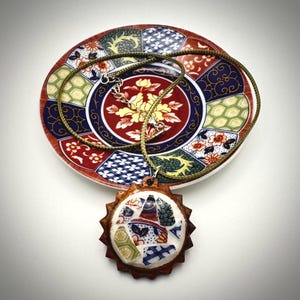 May include: A round pendant featuring a mosaic of broken ceramic shards in blue, red, white, and green, set within a dark brown wooden frame with a scalloped edge. The pendant hangs from a thin olive green cord, resting on a matching ceramic plate.