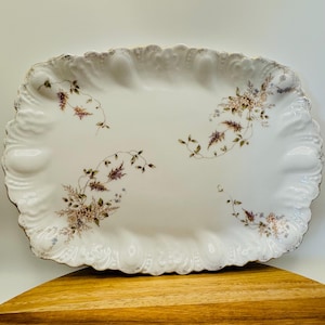 May include: An oval white porcelain serving platter featuring a scalloped edge with embossed detailing. The surface is decorated with delicate, scattered floral sprays in muted shades of brown, green, and soft purple, accented by gold trim.