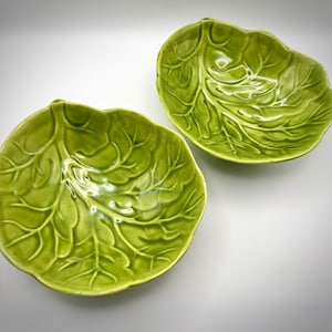 May include: Two matching bowls shaped like vibrant green cabbage leaves. Each bowl features a glossy finish with intricate, raised vein details that mimic the natural texture and organic form of a leafy vegetable, creating a realistic appearance.
