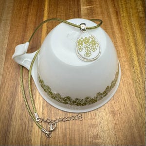 May include: A silver pendant necklace with a teardrop-shaped pendant featuring a floral design in shades of green. The pendant hangs on a green cord necklace, resting on a white bowl with a green floral pattern. The background is a wooden surface.