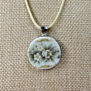May include: A round pendant necklace featuring a cream-colored ceramic disc with a hand-painted floral design in shades of gray, white, and yellow. The pendant is suspended from a light-colored cord and has a silver-toned metal bail.