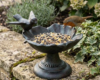 May include: A bronze-colored bird feeder with a pedestal base, filled with birdseed. A small bird perches on the edge, while a decorative bird sculpture is on the side. The feeder is placed on a stone wall, surrounded by greenery.