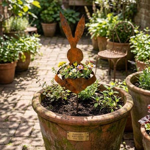 May include: A rusty metal rabbit-shaped planter holding colorful flowers. The planter is in a large terracotta pot filled with soil and greenery. The pot has a small tag that reads "GARDEN FAVORITE - EST. 2024."
