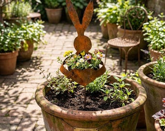 May include: A rusty metal rabbit-shaped planter holding colorful flowers. The planter is in a large terracotta pot filled with soil and greenery. The pot has a small tag that reads "GARDEN FAVORITE - EST. 2024."