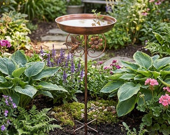May include: A copper-colored bird bath with a decorative hummingbird and flower design. The bird bath is filled with water and stands on a tall, slender metal pole with a three-pronged base. The bird bath is surrounded by lush green plants and flowers.