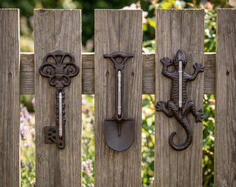 May include: Three decorative metal thermometers in the shape of a key, shovel, and lizard are mounted on a weathered wooden fence. The thermometers have red liquid inside, indicating the temperature. The background is blurred with greenery.