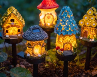 May include: Five solar-powered garden stake lights shaped like miniature houses. The houses feature varied designs including a green roof with 'Welcome' text, a red mushroom roof, a blue flower-covered roof, and a yellow sunflower-topped roof.