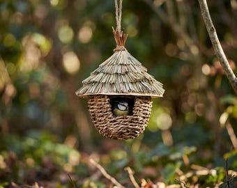 May include: A woven birdhouse with a thatched roof hangs from a tree branch. The birdhouse has a round, woven body and a small opening where a small bird is visible. The roof is made of straw and the birdhouse is suspended by a natural fiber rope.