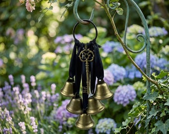 May include: A decorative garden bell chime with six small brass bells suspended from a black ribbon. A vintage key hangs in the center. The bells are hanging from a green metal hook in a garden setting.