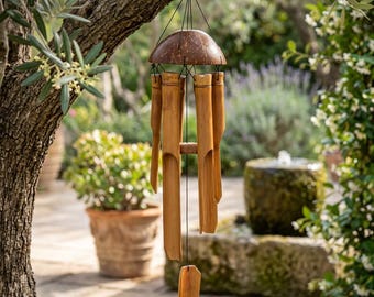 May include: A natural bamboo wind chime hanging from a tree branch. The chime features a coconut shell top and several bamboo tubes of varying lengths. The background shows a garden setting with greenery and a stone fountain.