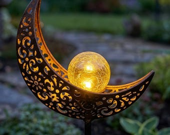 May include: A decorative solar-powered garden light featuring a crescent moon design with intricate cut-out patterns. The moon cradles a glowing yellow glass orb. The metal has a rustic, bronze finish, and the light is mounted on a black stake.