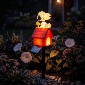 May include: A solar-powered garden light featuring a white Snoopy figure and a yellow Woodstock figure sitting on top of a red doghouse. The doghouse glows with an internal light, and a small solar panel is attached to the side of the black stake.