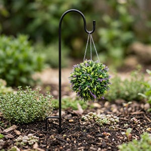 Peut inclure: Un piquet de jardin en métal noir avec un sommet incurvé et une chaîne suspendue supportant une plante artificielle sphérique. La plante présente des feuilles vertes et de petites fleurs violettes, sur un fond de jardin flou.