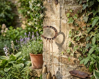 May include: A rustic horseshoe wind chime hanging on a brick wall. The wind chime features a brown horseshoe, jute rope, and three brass bells. The background includes lush greenery and a small terracotta pot.