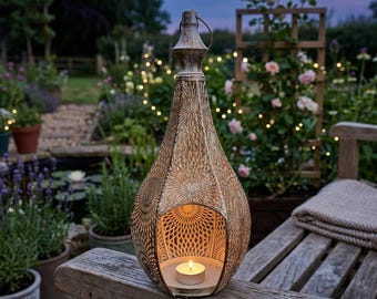 May include: A decorative, gold-colored metal lantern with a teardrop shape, intricate cut-out patterns, and a small candle inside. The lantern is placed on a wooden bench in a garden setting, with a blurred background of plants and lights.