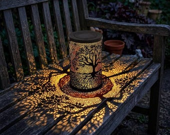 May include: A decorative solar-powered lantern with a tree design. The black lantern casts a tree-shaped shadow on the wooden bench. The lantern has a handle and is made of metal. The light is warm and inviting, perfect for outdoor decor.