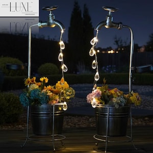 May include: Two garden planters with a water faucet design, featuring solar-powered lights. The planters are filled with flowers and have a vintage metal finish. The lights resemble water droplets, creating a charming nighttime display.