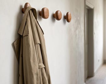 May include: A tan trench coat hangs on a row of four wooden wall hooks. The hooks are a light brown color and are mounted on a light-colored wall. The coat is made of a soft, flowing material.