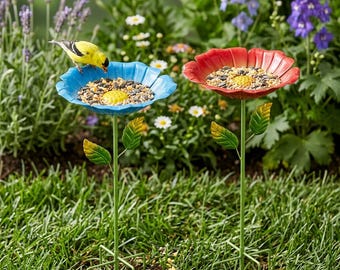 May include: Two flower-shaped bird feeders in a garden setting. One is blue, the other red, both filled with birdseed. A yellow bird perches on the blue feeder. Green stems and leaves support the flower-shaped bowls, set in a grassy area.