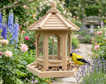 May include: A wooden bird feeder with a hexagonal base and a sloped roof hangs from a tree branch. The feeder has clear panels and is filled with birdseed. A yellow bird with black wings perches on the edge, eating. The background is a lush garden.