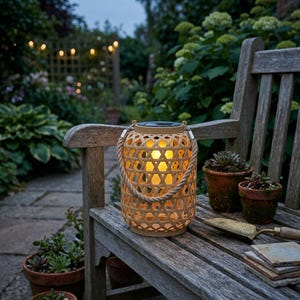 May include: A bamboo lantern with a woven design and a rope handle, illuminated by a warm yellow light, sits on a wooden bench. The background shows a garden with plants and fairy lights, creating a relaxing atmosphere.